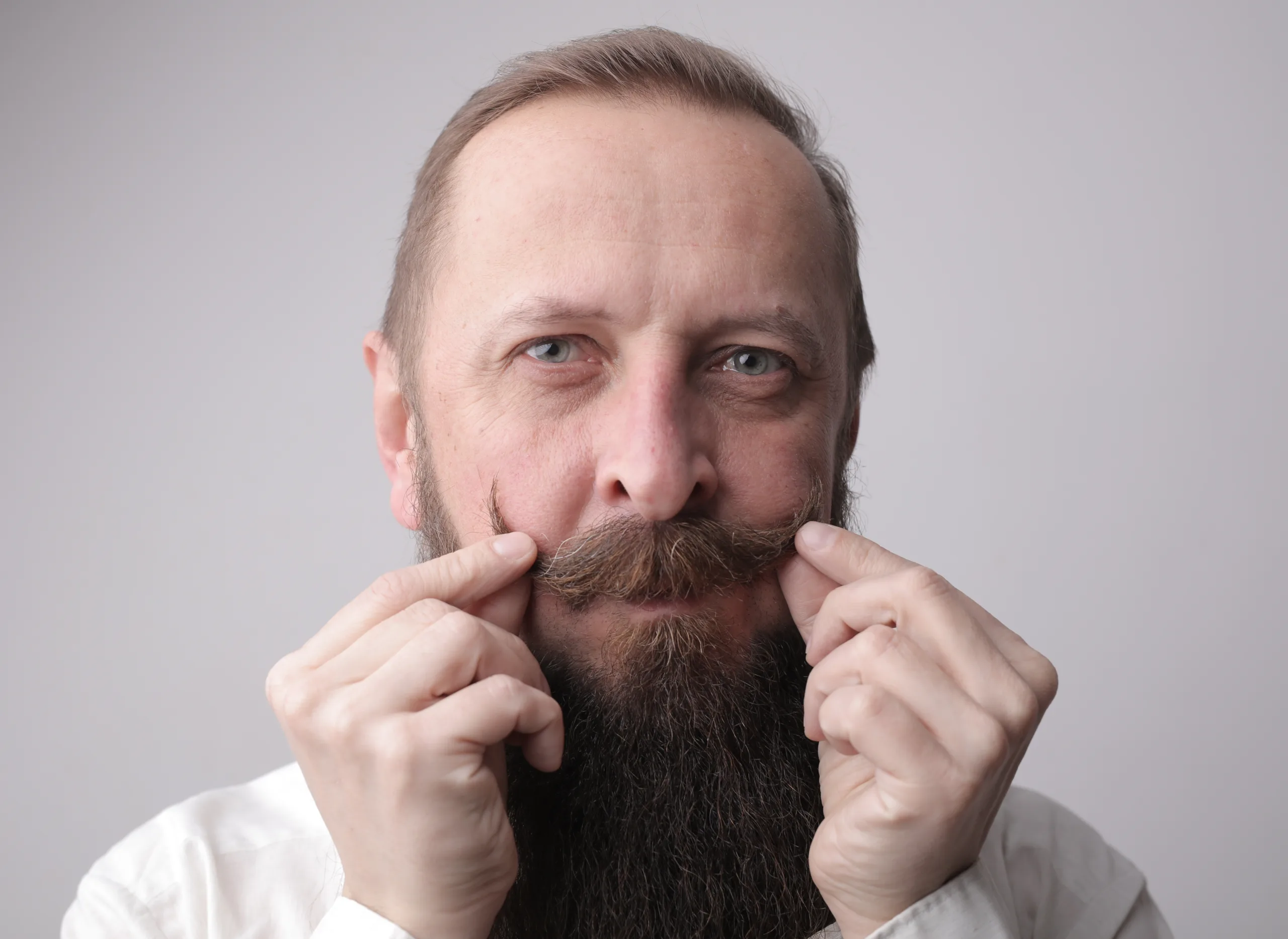 A man with a long beard and a mustache smiling while standing in front of a grey wall